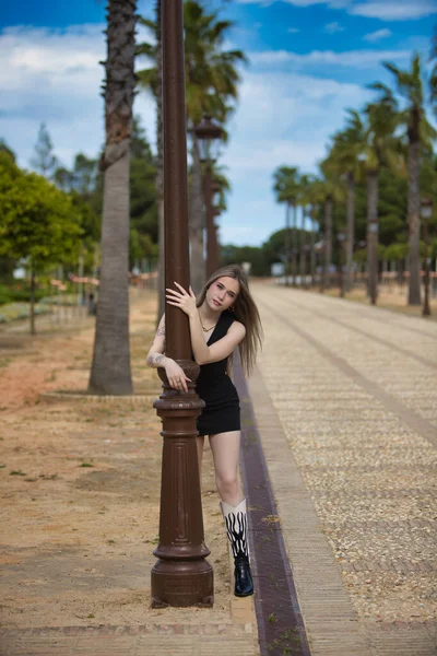 Siyah dövmeli, genç, güzel sarışın kadın palmiye ağaçları ve bahçeleri olan büyük bir caddede sokak lambasına sarılıyor. Kadın hafif bir gülümsemeyle utangaç görünüyor.