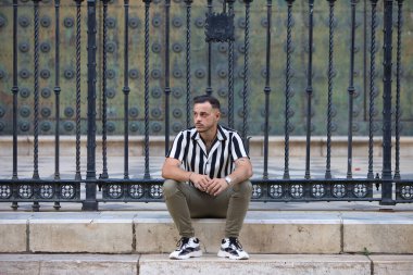 Genç, yakışıklı, siyah saçlı, çizgili gömlek ve kargo pantolonu giyen bir adam Seville, Endülüs 'teki Gotik katedralin önünde oturuyor. Adam şehirde tatilde.