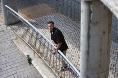 Sakallı, siyah tişörtlü ve bej kargo pantolonlu genç, yakışıklı bir adam, metal bir korkuluğa tutunuyor, rampadan aşağı indi ve yukarı bakıyor. Fotoğraf yukarıdan çekildi