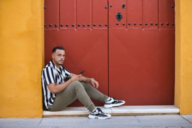 Çizgili gömlek ve kargo pantolonu giyen sakallı, genç, yakışıklı bir adam Seville, İspanya 'da kırmızı kapısı olan bir evin girişinde dinleniyor. Adam cep telefonunu kontrol ediyor.