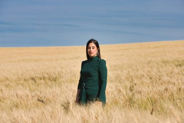 Yeşil giyinmiş, güzel, koyu renk saçlı Latin bir kız yaz günü kuru, sarı bir buğday tarlasının arasında duruyor. Arka planda, mavi gökyüzü