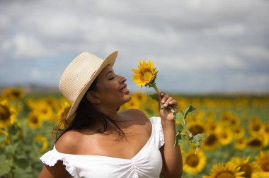 Beyaz gömlek ve hasır şapka giyip ayçiçeklerinin arasında duran koyu tenli genç bir Latin kadın çiftçinin portresi. Kız elinde bir çiçek tutuyor ve kokluyor ve ona bakıyor.