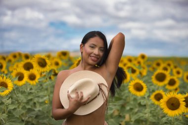 Genç, koyu tenli bir Latin kadın çiftçi, ayçiçeklerinin arasında beyaz bir bikini giyerek duruyor ve göğsünü elinde tuttuğu hasır bir şapkayla örtüyor. Kız doğada eğleniyor.