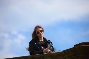 Güneş gözlüklü, siyah deri ceketli, çizmeli ve kısa etekli hoş bir sarışın kadın. Duvara yaslanmış havalı bir tavırla. Kadın gülümsüyor ve mutlu. Arka planda mavi gökyüzü