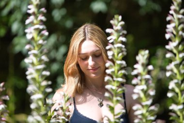 Güneşli ve sıcak bir günde kırsaldaki çiçeklerin arasında genç, güzel sarışın bir kadın. Kadın utangaç bir gülümsemeyle bakıyor.