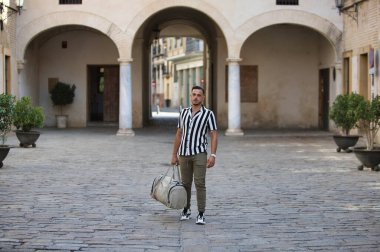 Genç, yakışıklı, siyah saçlı, çizgili tişörtlü ve kargo pantolonlu bir adam elinde bir seyahat çantası taşıyor ve eski bir şehrin sokaklarında yürüyor. Adam tatile geldi.