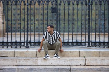 Genç, yakışıklı, siyah saçlı, çizgili gömlek ve kargo pantolonu giyen bir adam Seville, Endülüs 'teki Gotik katedralin önünde oturuyor. Adam şehirde tatilde.