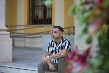 Çizgili gömlek ve kargo pantolonu giyen sakallı genç, yakışıklı bir adam bir binanın beyaz mermer merdiveninde oturuyor. Adam etrafa bakıyor.