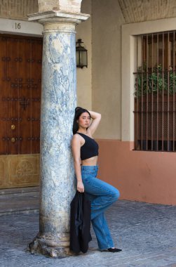 Genç, çekici, esmer, kot pantolon giyen ve siyah bluzlu bir kadın elinde bir blazer ceket tutuyor ve İspanya 'nın merkez bölgesinde bir caddede mermer bir sütuna yaslanıyor..