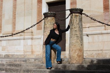 Koyu renk saçlı, kot pantolon giyen, siyah ceketli, taş sütunlar arasında duran ve Seville 'in merkezindeki eski bir binayı çevreleyen zincirlerle birlikte duran genç, çekici bir kadın.
