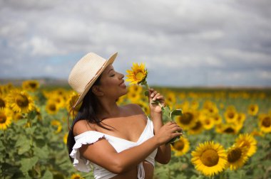 Beyaz gömlek ve hasır şapka giyip ayçiçeklerinin arasında duran koyu tenli genç bir Latin kadın çiftçinin portresi. Kız elinde bir çiçek tutuyor ve kokluyor ve ona bakıyor.