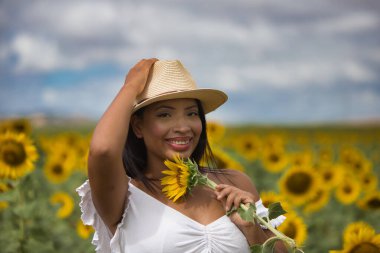 Beyaz gömlek ve hasır şapka giyip ayçiçeklerinin arasında duran koyu tenli genç bir Latin kadın çiftçinin portresi. Kız elinde bir çiçek tutuyor ve kokluyor ve ona bakıyor.