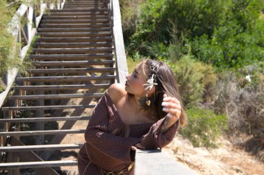 Saçında çiçekli, kahverengi elbiseli ve altın kemerli güzel bir genç kadın, bitki örtüsü arasında tahta bir merdivene yaslanıyor. Kadın farklı pozlar ve ifadeler veriyor.