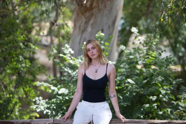 Genç, çekici, beyaz pantolon ve siyah pamuklu gömlek giyen sarışın bir kadın ormandaki nehir kenarındaki ahşap bir korkulukta oturuyor. Kadın dinleniyor ve bitki örtüsünün arasında dinleniyor.