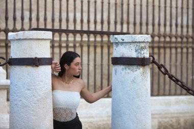 Beyaz dantelli bluz giyen, siyah şortlu ve deri çizmeli genç, çekici bir İspanyol kadın Seville Katedrali 'nin yanındaki iki taş sütun arasında çeşitli pozlar veriyor.
