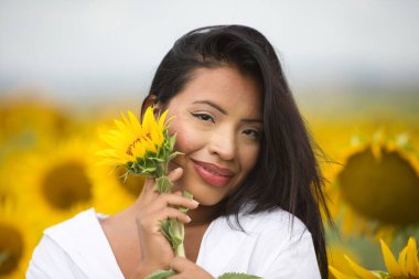 Beyaz gömlekli güzel bir melez Latin kadın onu yüzüne yaklaştırıyor ve bir ayçiçeğiyle flört ediyor. Kadın ayçiçeklerinin arasında mutlu ve güneşli yaz gününde gülümsüyor.