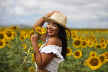 Beyaz gömlek ve hasır şapka giyip ayçiçeklerinin arasında duran koyu tenli genç bir Latin kadın çiftçinin portresi. Kız elinde bir çiçek tutuyor ve kokluyor ve ona bakıyor.