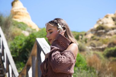 Saçında çiçekli, kahverengi elbiseli ve altın kemerli güzel bir genç kadın, bitki örtüsü arasında tahta bir merdivene yaslanıyor. Kadın farklı pozlar ve ifadeler veriyor.