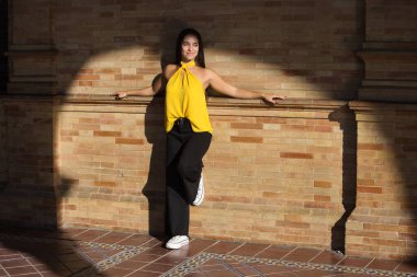 Genç, koyu saçlı, çekici Latin bir kadın sarı gömlek ve siyah pantolon giyiyor, kollarını açmış, mutlu görünüyor ve gülümsüyor.