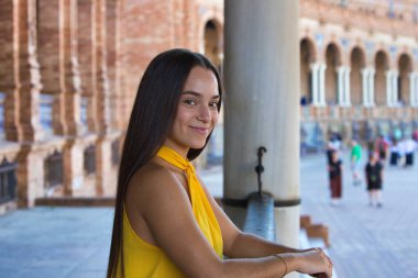 Genç, güzel, koyu renk saçlı, kırmızı giyinmiş, Sevilla Plaza de Espaa 'daki bir köprünün seramik korkuluklarına yaslanmış bir İspanyol kadın. Arka planda, anıtın bir kulesi.