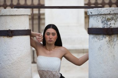 Beyaz dantelli bluz giyen, siyah şortlu ve deri çizmeli genç, çekici bir İspanyol kadın Seville Katedrali 'nin yanındaki iki taş sütun arasında çeşitli pozlar veriyor.