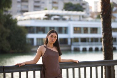 Seville nehri boyunca uzanan korkuluklara dayanan kahverengi bir elbise giyen genç ve güzel bir İspanyol kadının portresi. Kadın ciddi görünüyor. Arka planda nehir var.