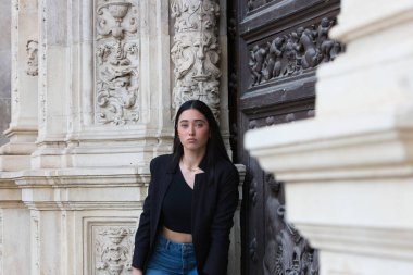 Gündelik kıyafetler giymiş ve siyah bir çanta taşıyan genç, çekici bir İspanyol kadın Plateresk tarzı Seville Belediyesi girişinin kapısına yaslanıyor.