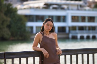 Seville nehri boyunca uzanan korkuluklara dayanan kahverengi bir elbise giyen genç ve güzel bir İspanyol kadının portresi. Kadın ciddi görünüyor. Arka planda nehir var.