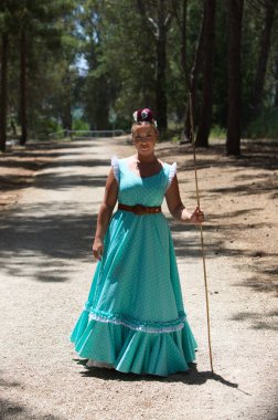 Geleneksel yeşil flamenko bir elbise giymiş güzel bir İspanyol kadın ve saçında bir çiçekle Hac yolculuğunda Endülüs 'ün çam ormanlarında bir çubuğa yaslanmış olarak yürüyor.