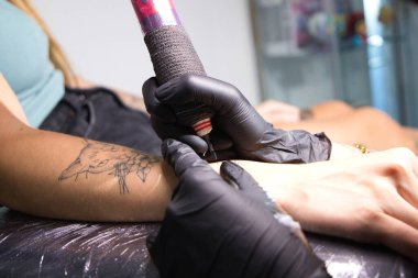 Genç kadın profesyonel dövme sanatçısı dövme makinesinin iğnesini kullanarak cildine siyah mürekkep sokuyor.