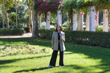 Siyah saçlı, genç ve çekici bir İspanyol kadın parkta farklı pozlar veriyor. Siyah kot pantolon, boynunda atkısı olan siyah bir tişört ve bej bir ceket giyiyor.