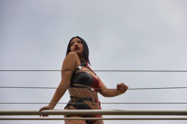 Güzel, siyah saçlı, zombi gibi giyinmiş, kırmızı ve siyah deri bir bluz ve şort giyen siyah balıkçı çoraplı ve siyah çizmeli bir Latin Amerikalı kadın. Kadın merdiven korkuluklarına yaslanmış.