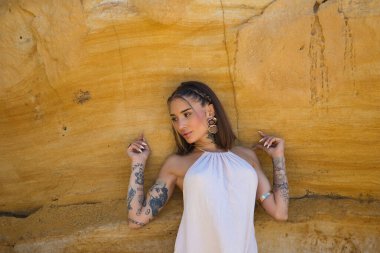 Makyaj yapan güzel bir genç kadın, bej bir elbise ve deniz kenarındaki bir uçurum duvarına dayanmış altın mücevherler. Kadın farklı pozlar ve ifadeler veriyor. Güzellik ve moda kavramı