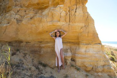 Makyaj yapan güzel bir genç kadın, bej bir elbise ve deniz kenarındaki bir uçurum duvarına dayanmış altın mücevherler. Kadın farklı pozlar ve ifadeler veriyor. Güzellik ve moda kavramı