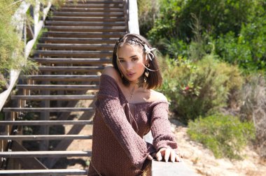 Saçında çiçekli, kahverengi elbiseli ve altın kemerli güzel bir genç kadın, bitki örtüsü arasında tahta bir merdivene yaslanıyor. Kadın farklı pozlar ve ifadeler veriyor.