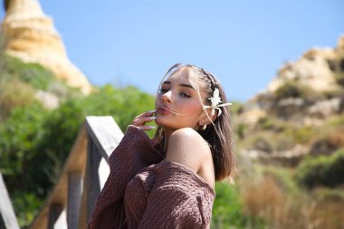 Saçında çiçekli, kahverengi elbiseli ve altın kemerli güzel bir genç kadın, bitki örtüsü arasında tahta bir merdivene yaslanıyor. Kadın farklı pozlar ve ifadeler veriyor.