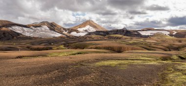 Rugged Icelandic peaks with green slopes, snow crevices, and dramatic volcanic terrain under overcast sky.