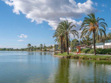 Sakin, geniş bir göl manzarası palmiye ağaçları, yeşil bir çimen ve bulutlu bir gökyüzünün altındaki tatil köyleri..