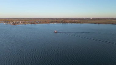 Küçük bir teknenin hareket ettiği sakin bir göl manzarası ve arkasında tekneler olan bir marina..