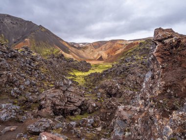 Öndeki yosunlu, karanlık lav kayaları renkli rhyolite dağları arasında dar bir vadiye çıkar..
