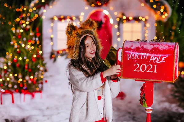 A beautiful girl sends a letter to Santa in a red mailbox