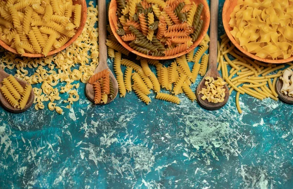 Tahta kaşıklarda çeşitli kuru makarna karışımları. Mavi arka planda kuru makarna. Kopyalama alanı olan üst görünüm. Diyet ve yemek konsepti.