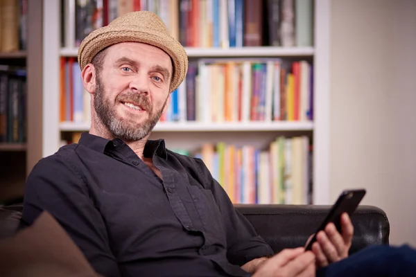 Hipster con sombrero sentado en su sala de estar con un teléfono móvil — Foto de Stock