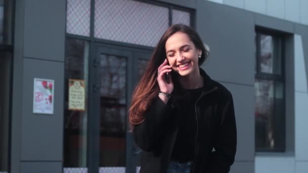 Дуже красива дівчина розмовляє по телефону і посміхається. Дівчина стоячи в місті, поруч з рестораном . — стокове відео