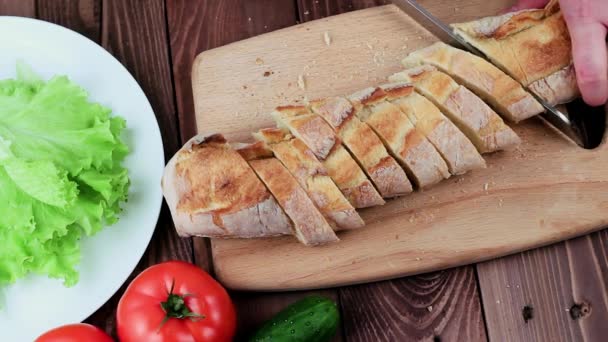 Cook snijdt Frans stokbrood. Kruimels opgestapeld op een bord. Ook op de tafel zijn tomaten, komkommers en sla. Top View. — Stockvideo