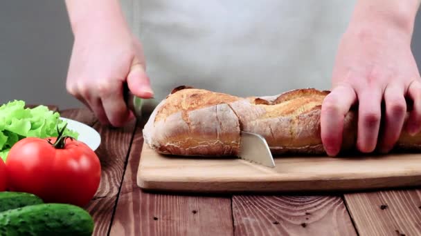 Köchin schneidet frisches französisches Baguette. Krümel stapeln sich auf einem Teller. Auch Tomaten, Gurken und Salat stehen auf dem Tisch. — Stockvideo