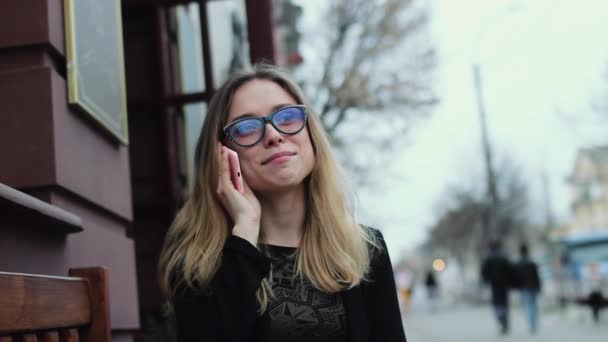 Bella ragazza con gli occhiali, parlando al telefono mentre seduto su una panchina in città. La ragazza sorride. Lei è molto felice e amichevole . — Video Stock