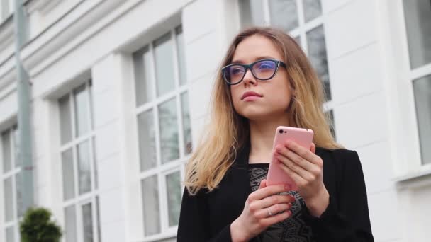 Schönes und nettes Mädchen mit Brille, das in der Nähe der Universität wartet. der Student hält das Telefon in der Hand und glättet die Haare. — Stockvideo