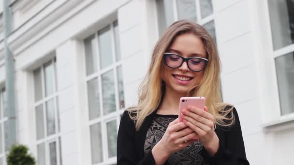 Velmi krásná a atraktivní dívka s brýlemi píše zprávu na telefon stojící Outside the úřadu. Žena má šťastný vzhled, usmívá se. — Stock video