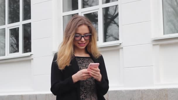 Дуже красива і мила дівчина в окулярах вводить повідомлення на телефон поки стоїть біля університету. Жінка має щасливий погляд, вона посміхається. — стокове відео